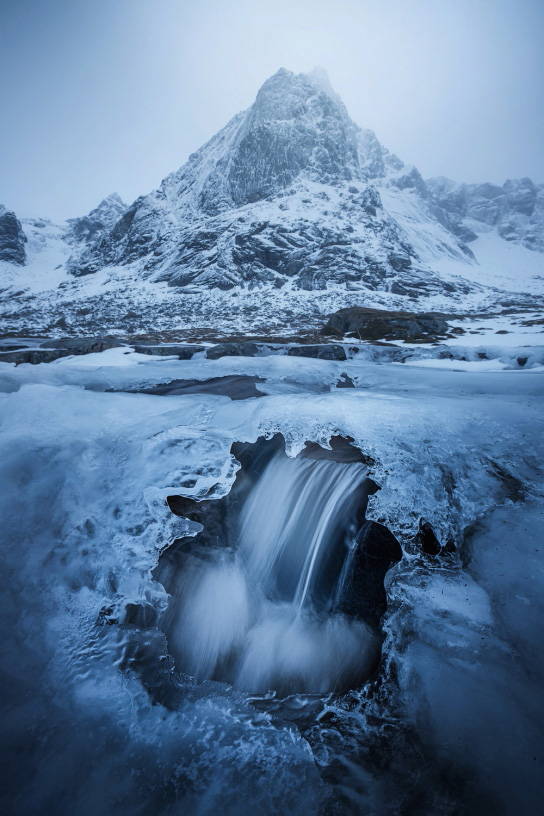 Vertical shot of waterfall beneath the ice in front of mountain in Norway